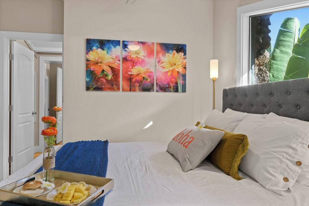 A hotel room with a bed, a painting of flowers, a window with a view, and a tray of food on the nightstand.