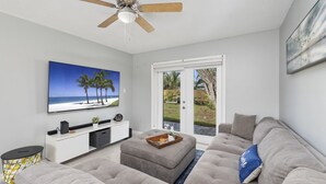 Ein modernes Wohnzimmer mit einem großen Flachbildfernseher, einem Deckenventilator, einem grauen Sofa und einer Glastür, die auf eine Terrasse führt.