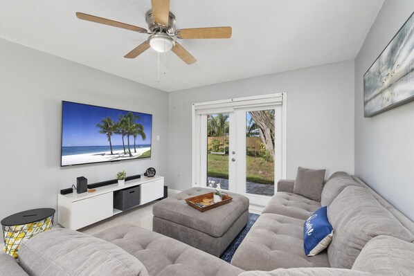 A modern living room with a large flat-screen TV, a ceiling fan, a grey sofa, and a sliding glass door leading to a patio. - Beautiful Island Home to relax! (Fort Pierce)