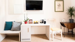 Un espace de vie compact comprenant un bureau blanc, une chaise, un petit meuble, un téléviseur fixé au mur et une plante en pot.