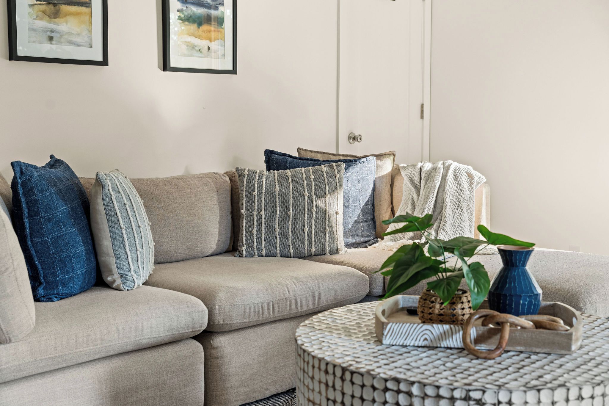 Un salon avec un canapé d’angle beige, de nombreux coussins, une table basse ronde agrémentée d’objets décoratifs et deux œuvres d’art encadrées accrochées au mur.