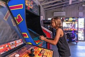 Una persona juega videojuegos en un salón recreativo con máquinas coloridas y un baúl de juguetes al fondo.