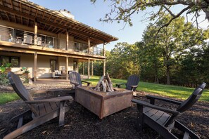 Un espacio al aire libre moderno con sillones Adirondack y un brasero, rodeado por una terraza de madera y un árbol grande.