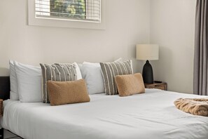 Una habitación de hotel con cama, almohadas, una mesita de noche con lámpara y una ventana con persianas.