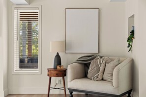 Una habitación con ventana y persianas, una lámpara en una mesita auxiliar, un sofá con cojines y una obra de arte enmarcada en la pared.