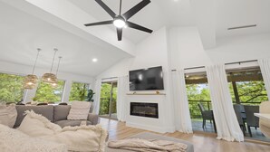 Un salón espacioso con ventilador de techo, televisor de pantalla plana, chimenea y ventanas amplias que dan a zonas verdes.