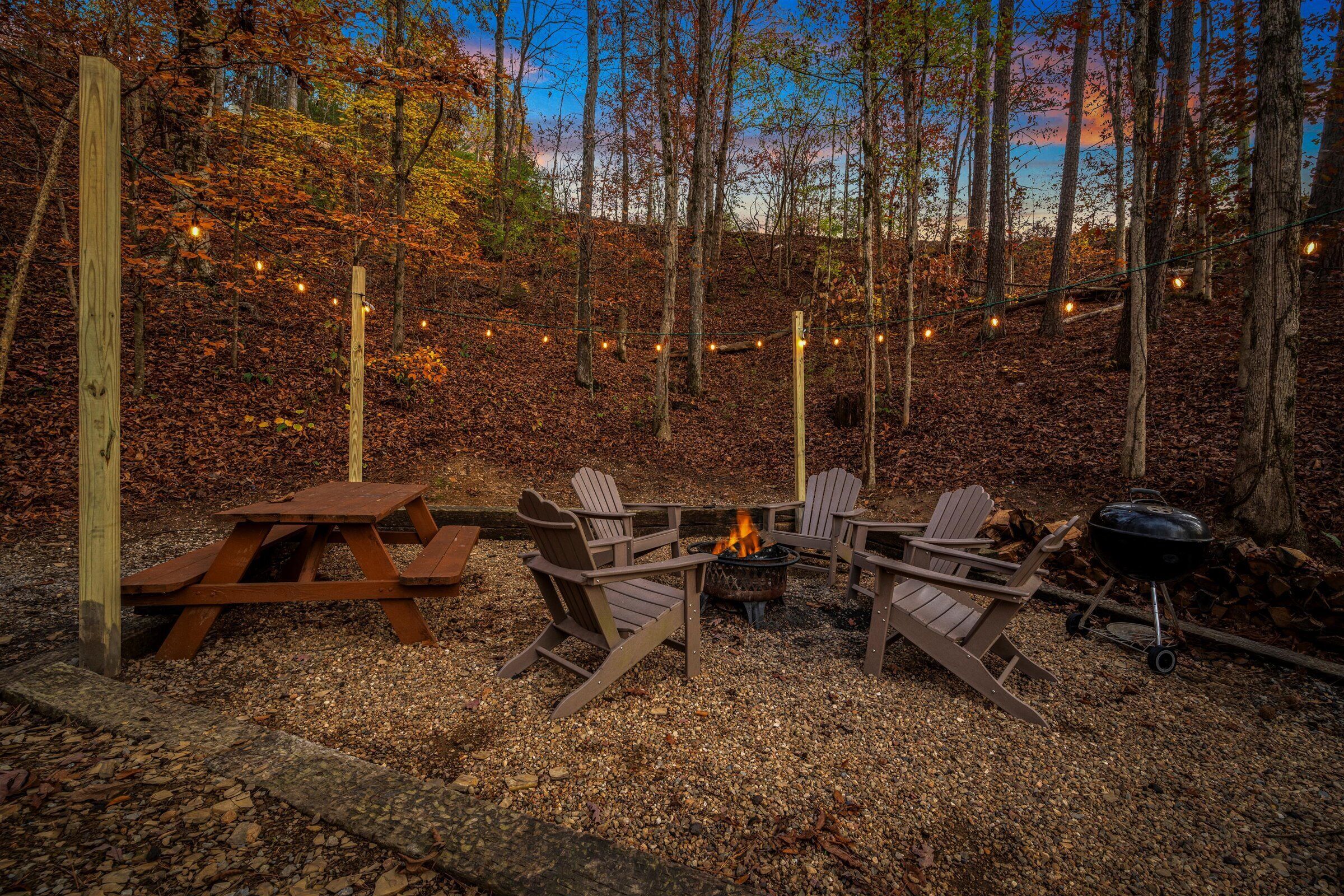Un espace extérieur rustique aménagé avec un foyer, des chaises en bois et une table de pique-nique, le tout entouré d’une forêt.