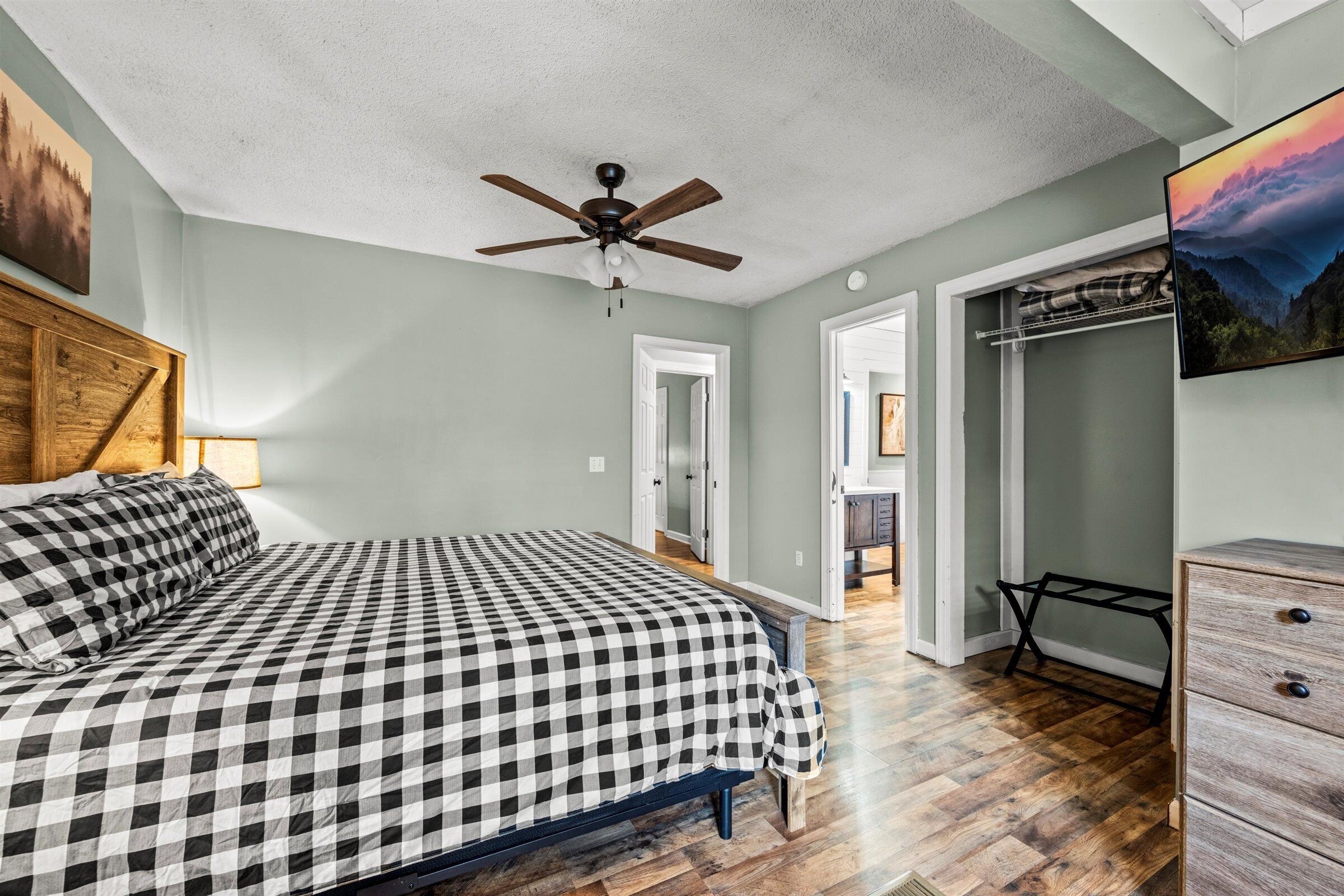 A bedroom with a checkered bedspread, a ceiling fan, a wooden dresser, and a painting on the wall.