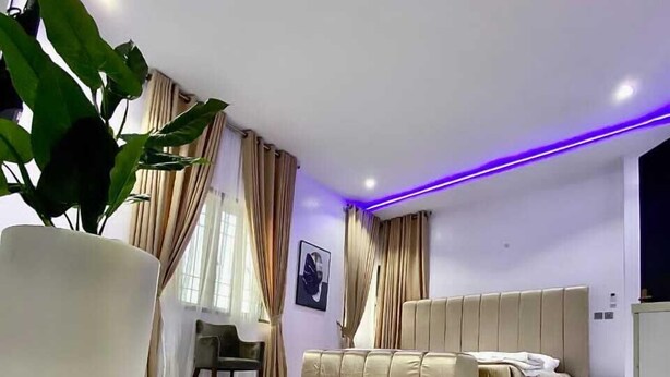 A modern living room with a large white sofa, a dining area with a chair, and a large potted plant. - Harrington court (Lekki)