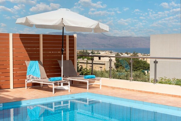 A poolside area with a view of the sea and mountains, wooden paneling, and two lounge chairs with blue cushions. - Villa Flora - Villa close to the seaside with private pool in Crete (Pýrgos Psilonérou)