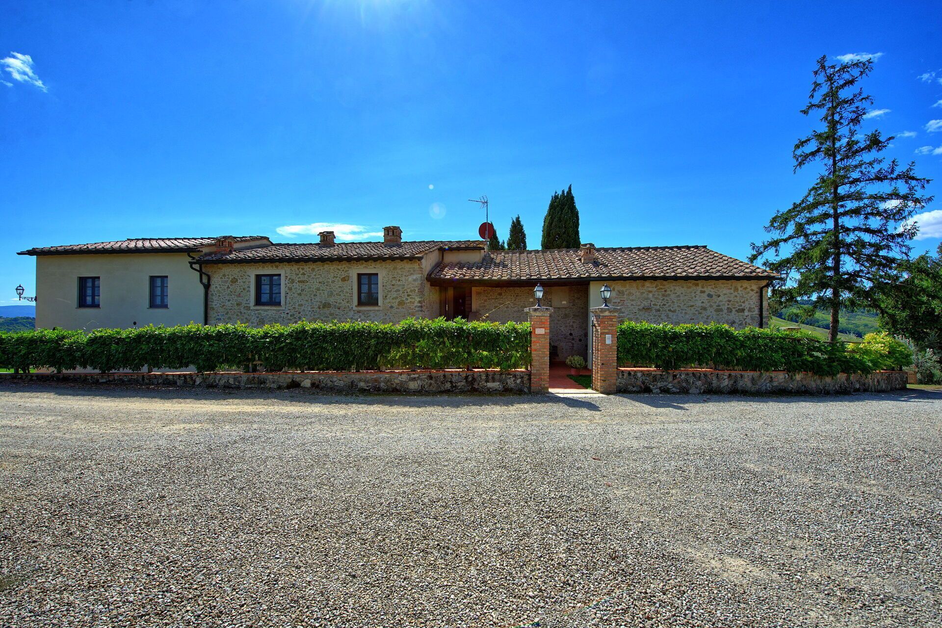A stone building with a tiled roof, a gravel driveway, and a hedge in front.