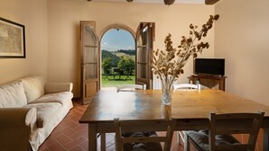 Un coin repas avec une table et des chaises en bois, un vase contenant des fleurs séchées, un téléviseur posé sur un meuble, et une vue sur un paysage verdoyant par une porte ouverte.