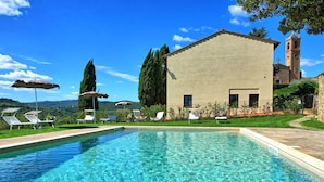 A swimming pool with lounge chairs and umbrellas, a building with a tower, and a view of hills. - Camino, Montauto - Holiday apartment  with pool close to San Gimignano (Montauto)