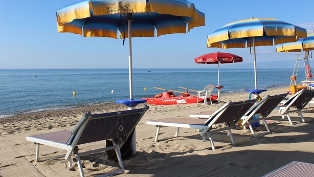 Une plage bordée de rangées de chaises longues et de parasols, un poste de surveillance pour les sauveteurs et un petit bateau sur l’eau.
