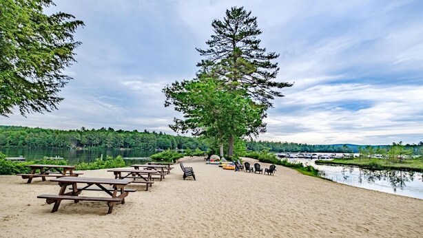 A sandy beach with picnic tables and chairs, a large tree, and a calm lake in the background. - Lakes & Mountain Experience! 4 Seasons Getaway -- 3 Bed 1.5 Bath Cozy Cabin (Bridgton)