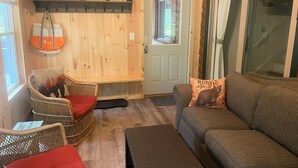 A cozy room with a wooden interior, a grey sofa, wicker chairs, a bench, and a door with a window. - Lakes & Mountain Experience! 4 Seasons Getaway -- 3 Bed 1.5 Bath Cozy Cabin (Bridgton)