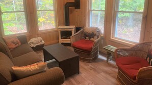 A cozy cabin interior with a fireplace, wicker furniture, and large windows offering views of the forest. - Lakes & Mountain Experience! 4 Seasons Getaway -- 3 Bed 1.5 Bath Cozy Cabin (Bridgton)