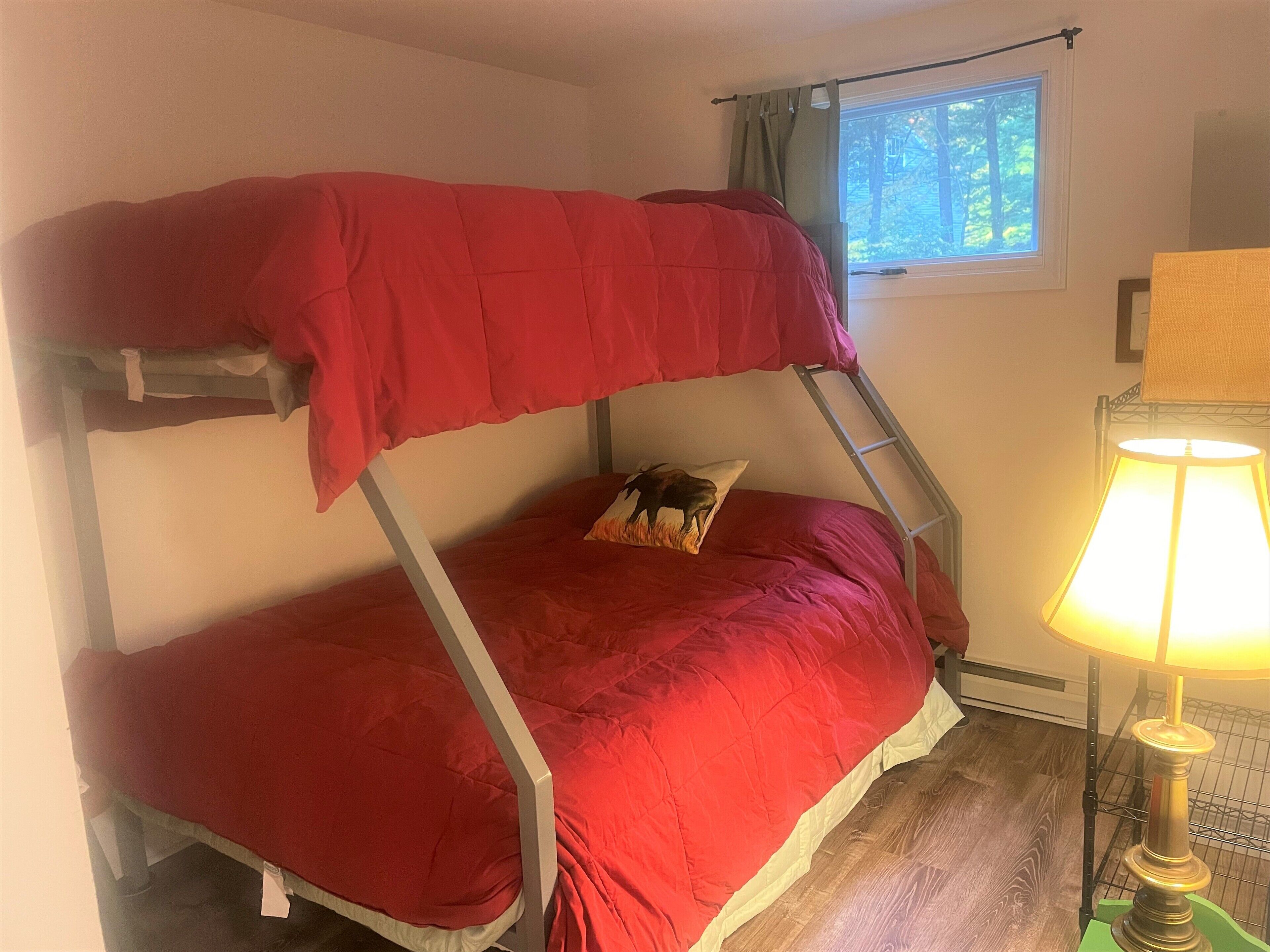 Una cama litera con un colchón rojo y una almohada con un diseño de búfalo. Se ve una ventana con vistas a árboles y una lámpara en el lado derecho.