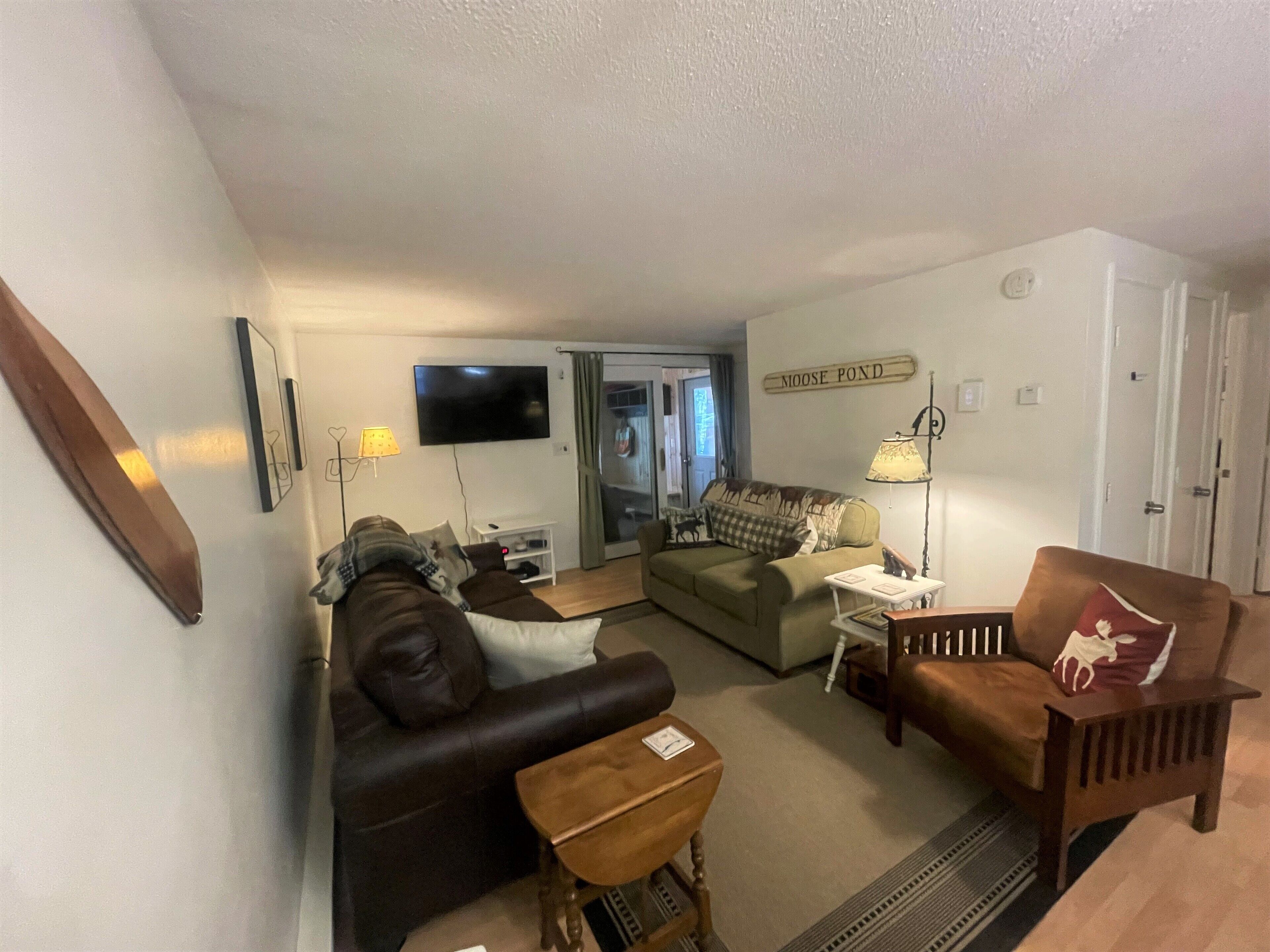 A cozy living room with a leather sofa, a wooden armchair, and a small table. A television is mounted on the wall, and a sign reads "NOOSE POND".