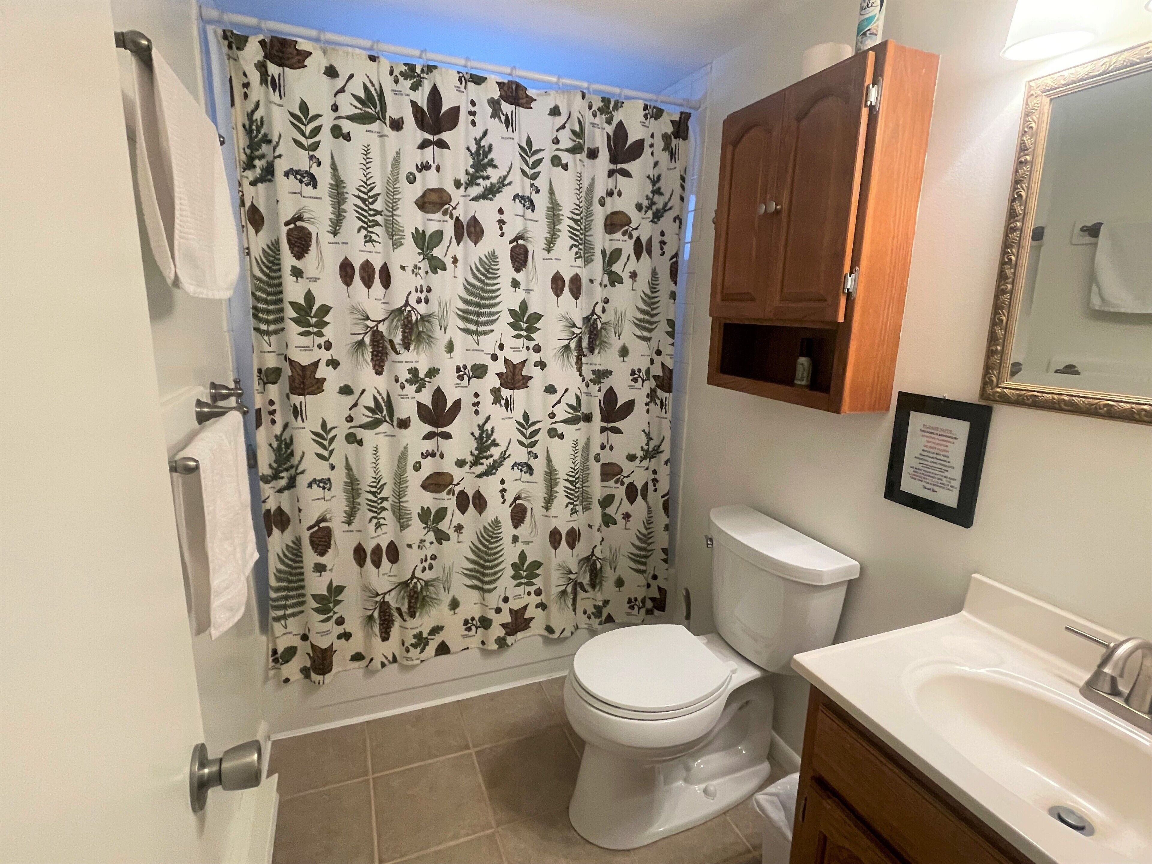 Baño con cortina de ducha estampada con hojas, inodoro, lavamanos y un mueble de madera sobre el lavamanos.