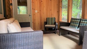 A cozy room with a wicker sofa, a chair with a striped pillow, and a view of trees through large windows. - Lakes & Mountain Experience! 4 Seasons Getaway -- 3 Bed 1.5 Bath Cozy Cabin (Bridgton)