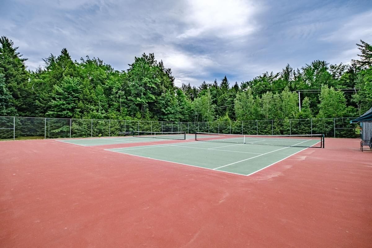 An outdoor tennis court with a green and red surface, surrounded by a metal fence and trees.