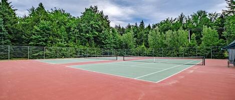 An outdoor tennis court with a green and red surface, surrounded by a metal fence and trees.