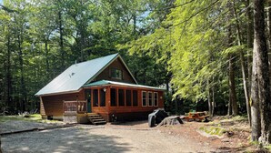 A rustic cabin with a green roof, surrounded by dense forest. - Lakes & Mountain Experience! 4 Seasons Getaway -- 3 Bed 1.5 Bath Cozy Cabin (Bridgton)