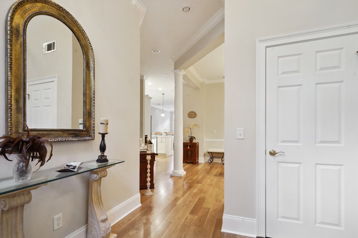 A hallway with a mirror, a glass shelf with a vase, a door, and a view into a room with a bench and a plant.