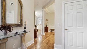 A hallway with a mirror, a glass shelf with a vase, a door, and a view into a room with a bench and a plant. - 10 Palm Villas Court (St Simons Island)