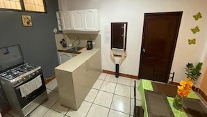 Cozinha compacta com fogão a gás, pia e uma pequena ilha. Há uma janela com persianas, uma porta e uma prateleira na parede.