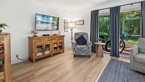 A living room with a wooden cabinet, a patterned armchair, a small table, and a large window with curtains. - Free bikes/pools/tennis/fitness Walk 2 pier/village shops & restaurants! (St Simons Island)