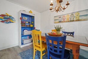 Ein Essbereich mit einem Holztisch, blauen Stühlen, einem gelben Stuhl, einem blauen Schrank mit Stauraum, einer Pendelleuchte und einem Gemälde von Strandhäusern.