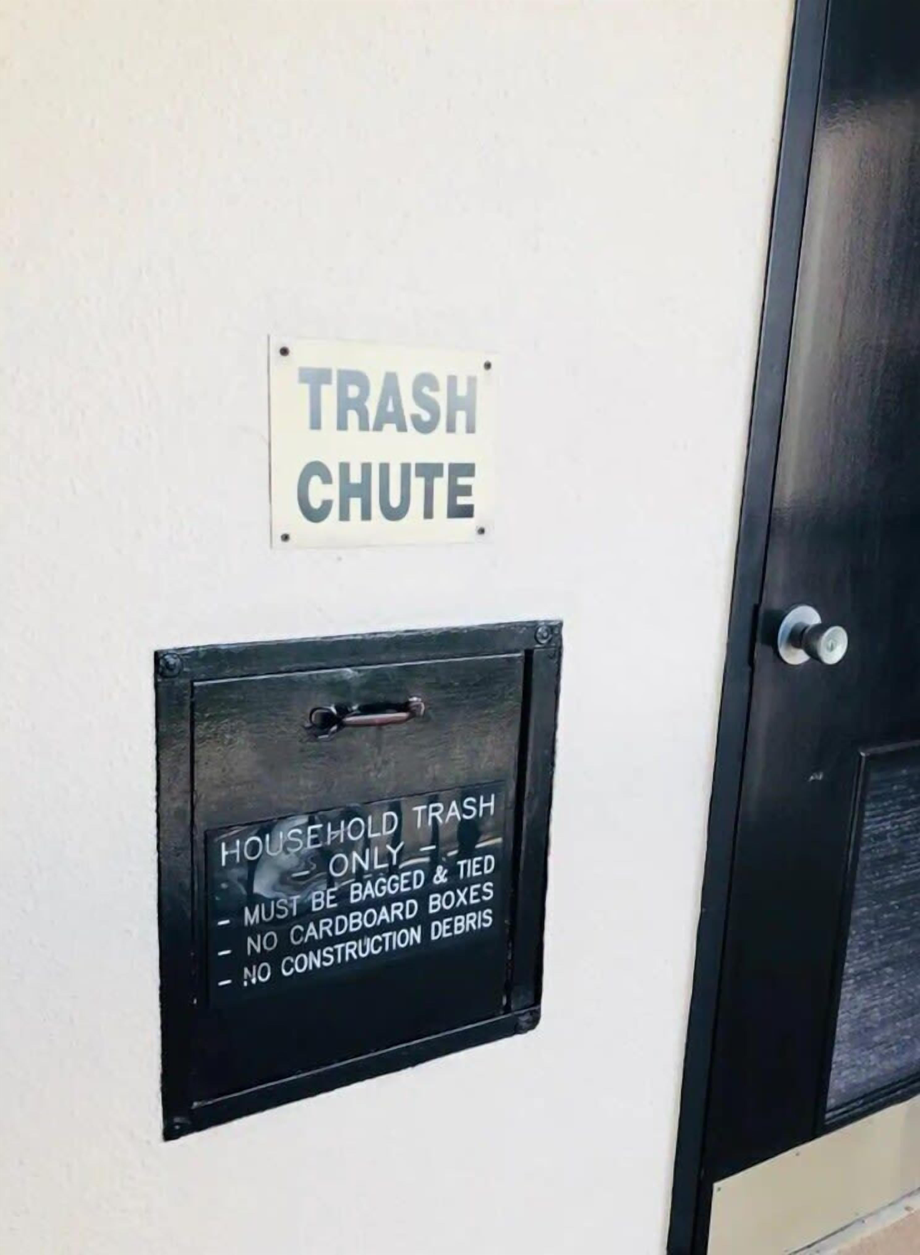 A trash chute with a sign above it that reads "TRASH CHUTE" and another sign below it that reads "HOUSEHOLD TRASH ONLY - MUST BE BAGGED & TIED - NO CARDBOARD BOXES - NO CONSTRUCTION DEBRIS".