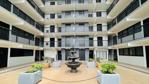 Un edificio de varios pisos con un patio central que tiene una fuente, rodeado de plantas en macetas y una barrera de cadena.