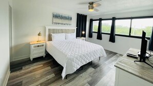Un dormitorio con cama, mesita de noche, escritorio con ordenador y ventana con cortinas.