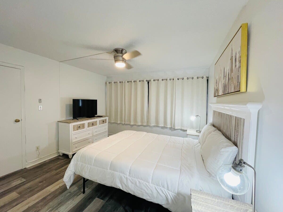 A bedroom with a bed, a dresser, a television, a ceiling fan, and a window with curtains.