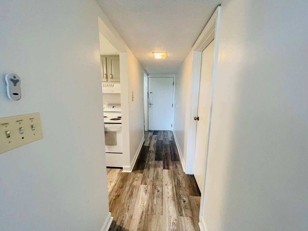 Un couloir étroit au plancher en bois, une porte blanche au fond et un mur blanc avec un interrupteur et des prises électriques.