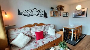 En hyggelig stue med en træsofa, et lille bord med en plante og en vægmonteret reol med pynteting.