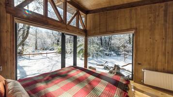 A wooden cabin room with a bed, large windows, and a view of a snowy forest.