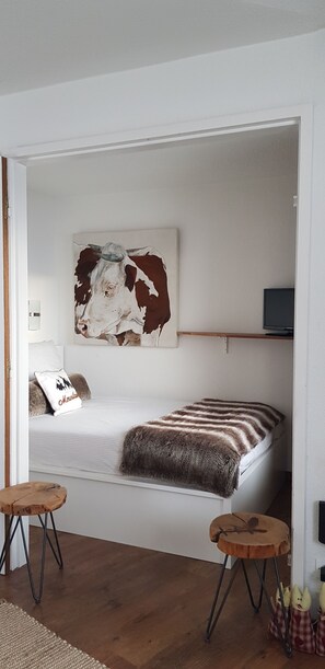 Un dormitorio con cama, televisor montado, un cuadro y un taburete pequeño.