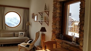 A cozy room with a stone wall, a round window with a snowy mountain view, a sofa with a pillow, a wooden chair, and a small table with a lamp. - Studio 4 P ideally located with magnificent view (Morzine)