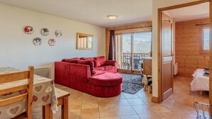 A cozy living room with a red sofa, a wooden dining table, and a view of mountains through the glass door. - Floor -2, view mountain, swimming pool, balcony, tv, ski locker, 70m², Les Arcs (Bourg-Saint-Maurice)