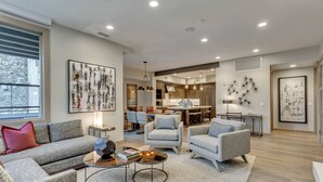 A modern living room with a grey sofa, armchairs, and a coffee table. There is a dining area with a table and chairs, and a kitchen in the background.