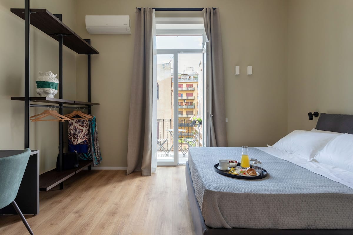 Une chambre d’hôtel comprenant un lit, un bureau, une chaise, un porte-vêtements et un balcon avec vue sur les bâtiments.