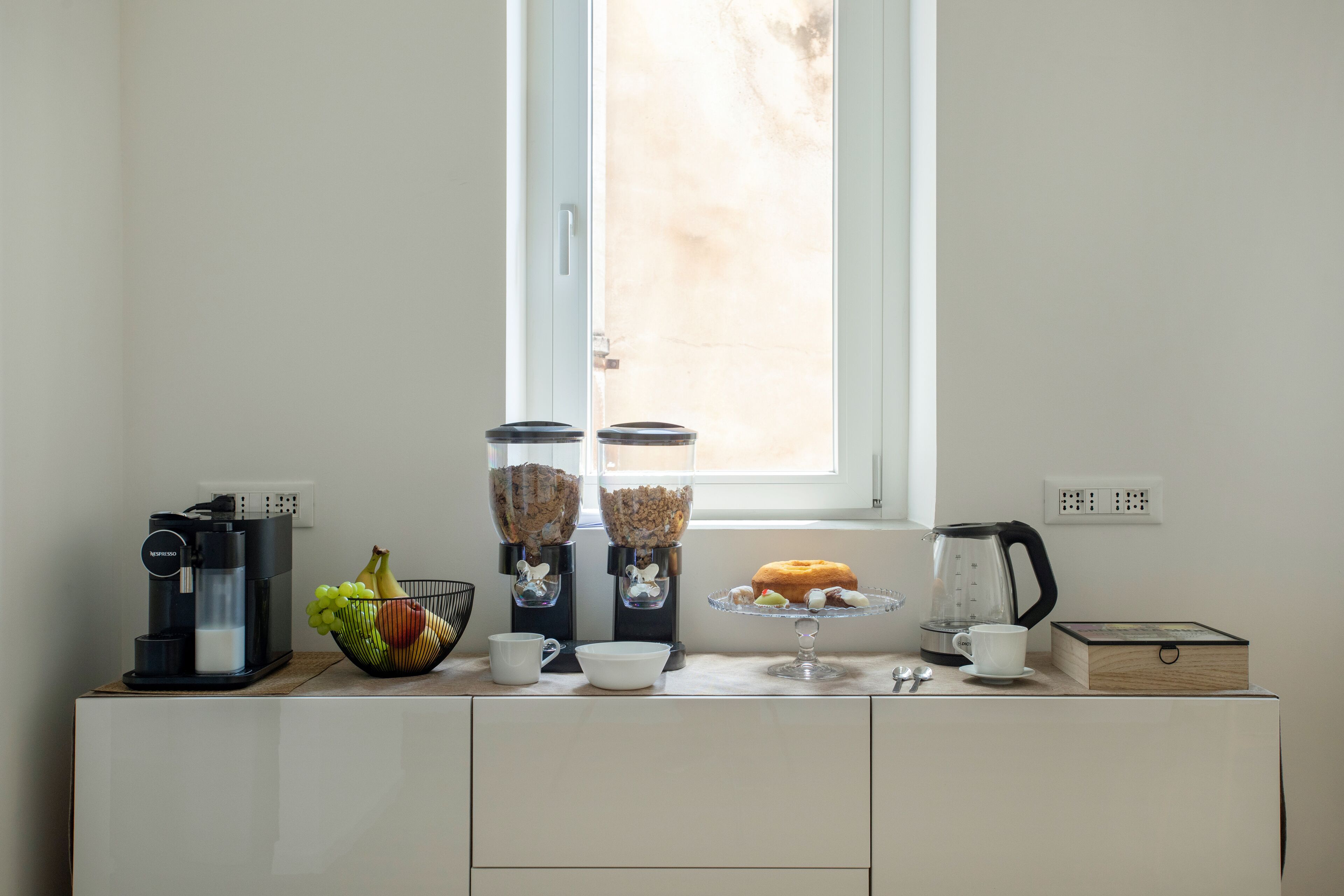 A kitchen counter with a coffee maker, two coffee dispensers, a fruit bowl, a glass cake stand with a donut, and a coffee pot.