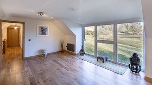 A spacious room with wooden flooring, large windows offering a view of a grassy landscape, a door leading to another room, and a small statue on a stool. - The Stables - near St Andrews Sleeps 10 (Fife)