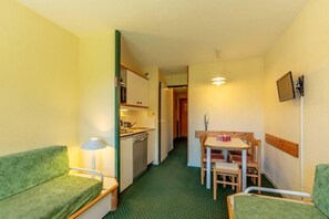 A compact hotel room with a kitchenette, a dining area with a table and chairs, and a green patterned sofa.