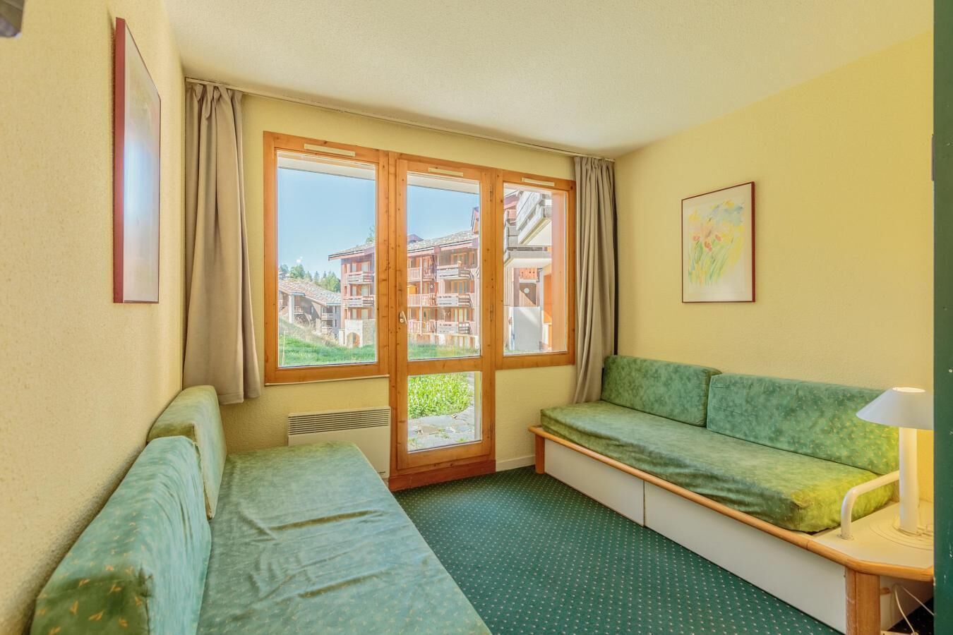 Une pièce avec un canapé vert, une petite table avec une lampe et une fenêtre donnant sur des immeubles.
