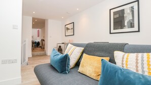 Uma sala de estar moderna com um sofá cinzento, almofadas azuis e amarelas, um quadro emoldurado na parede e uma escadaria que dá para outro cômodo.